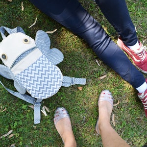 May include: A gray and white chevron patterned backpack with a white fabric face and two large black eyes. The backpack is lying on green grass.