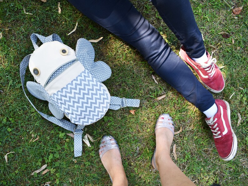 May include: A gray and white chevron patterned backpack with a white fabric face and two large black eyes. The backpack is lying on green grass.