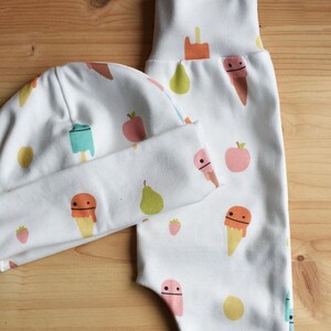 May include: A white baby hat and pants set with a fun ice cream and fruit print. The hat has a fold-over brim and the pants have a fold-over waistband.