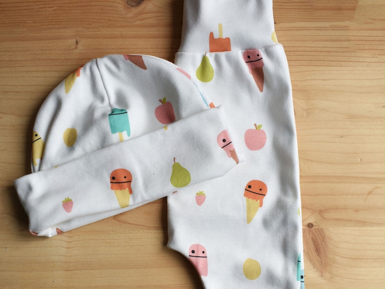 May include: A white baby hat and pants set with a fun ice cream and fruit print. The hat has a fold-over brim and the pants have a fold-over waistband.