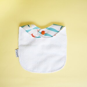 May include: White terry cloth baby bib with a blue and white patterned trim featuring orange crabs. The bib has a snap closure and the text "Zeila" is printed on the bib.
