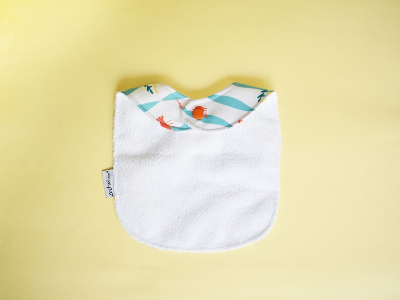 May include: White terry cloth baby bib with a blue and white patterned trim featuring orange crabs. The bib has a snap closure and the text "Zeila" is printed on the bib.