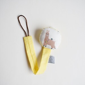 Puede incluir: Sujeta chupetes para beb&eacute; amarillo y blanco con un dise&ntilde;o de llama. El clip tiene un lazo marr&oacute;n en la parte superior y una etiqueta blanca con texto negro.