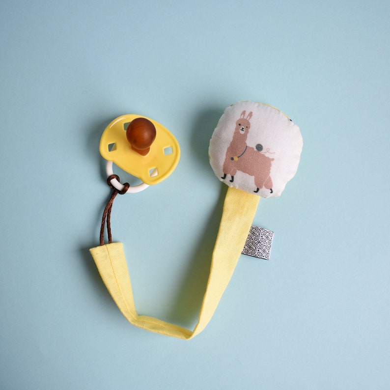 Puede incluir: Un clip para chupete amarillo con un chupete marr&oacute;n y amarillo adjunto. El clip tiene una llama de tela blanca con manchas marrones y una cuerda amarilla adjunta.