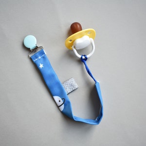 May include: A baby pacifier with a blue fabric strap featuring a space-themed pattern of planets and stars. The pacifier has a yellow shield, a brown nipple, and a white handle. A light blue clip is attached to the strap.
