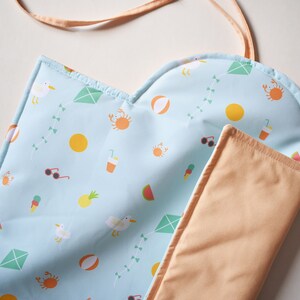 May include: A light blue beach mat with a summer pattern of kites, crabs, and ice cream cones. It has a peach-coloured lining and a matching strap. Ideal for a day at the beach or park. The mat is approximately 150 cm long.