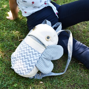 May include: A gray and white patterned backpack with a zippered pocket and two large, rounded, gray wings. The backpack has two large, brown buttons on the front.