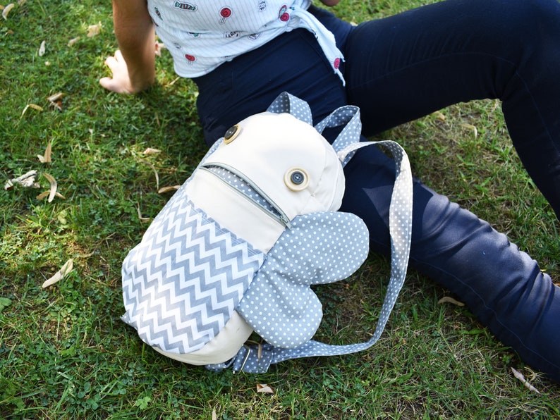 May include: A gray and white patterned backpack with a zippered pocket and two large, rounded, gray wings. The backpack has two large, brown buttons on the front.