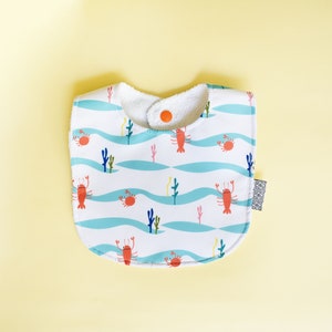 May include: A white baby bib with a blue and white wave pattern and red lobsters and crabs. The bib has a white terry cloth lining and a snap closure.
