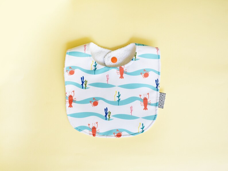 May include: A white baby bib with a blue and white wave pattern and red lobsters and crabs. The bib has a white terry cloth lining and a snap closure.