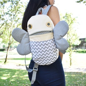 May include: A gray and white patterned backpack with large gray wings and two button eyes. The backpack has a zippered pocket and a chevron pattern on the front.