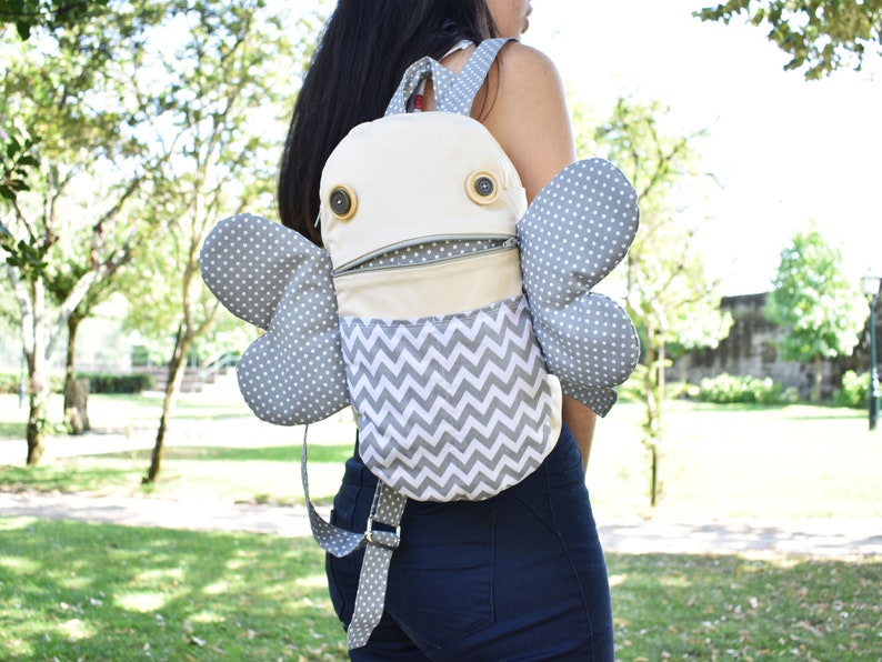 May include: A gray and white patterned backpack with large gray wings and two button eyes. The backpack has a zippered pocket and a chevron pattern on the front.