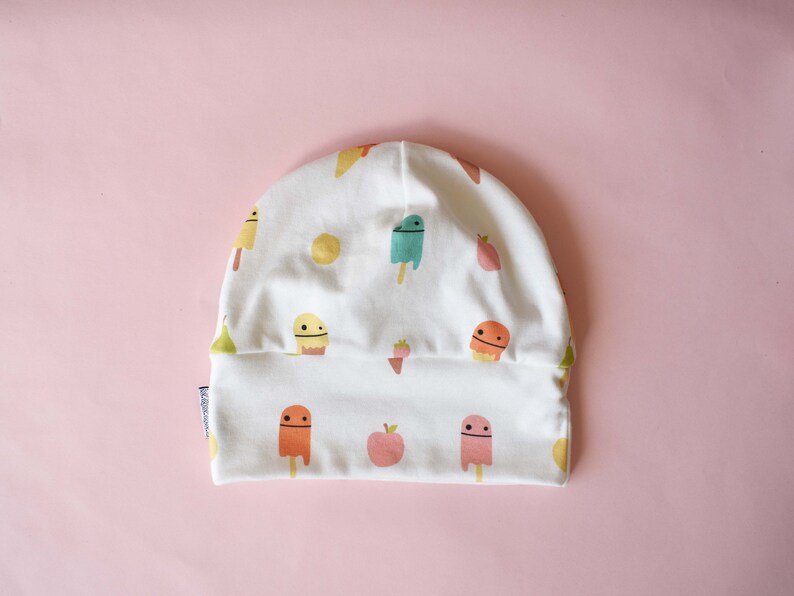 May include: White cotton beanie with a colorful pattern of ice cream cones and fruit. The ice cream cones have smiling faces.