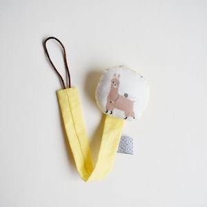 Puede incluir: Sujeta chupetes para beb&eacute; amarillo y blanco con un dise&ntilde;o de llama. El clip tiene un lazo marr&oacute;n en la parte superior y una etiqueta blanca con texto negro.