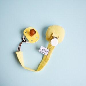 Puede incluir: Clip para chupete amarillo con un porta chupete amarillo y un clip blanco. El clip tiene una etiqueta blanca con el texto "zezling!" y la direcci&oacute;n del sitio web "www.zezling.com".