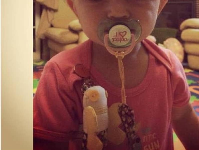 Puede incluir: Un ni&ntilde;o peque&ntilde;o con una camisa rosa con un chupete sujeto a un juguete. El chupete tiene un dise&ntilde;o rosa y blanco con el texto "I love papa" en &eacute;l. El juguete es un b&uacute;ho de peluche con un ojo de bot&oacute;n.