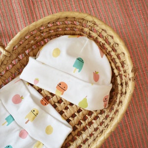 May include: A white baby hat and matching pants with a colorful ice cream and fruit pattern. The hat and pants are in a woven basket.