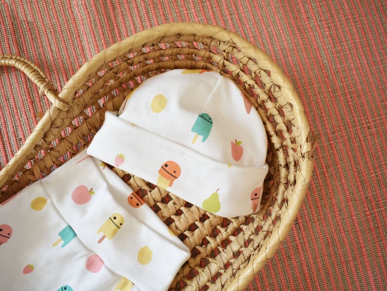 May include: A white baby hat and matching pants with a fun pattern of colorful popsicles, lemons, and strawberries.
