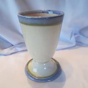 May include: A white ceramic vase with a blue and green band around the top and bottom. The vase has a flared top and a round base.