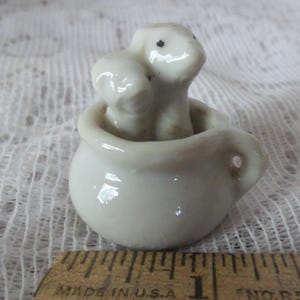 May include: White ceramic figurine of two dogs sitting in a white ceramic pot with a handle. The dogs have black eyes and are about 1 inch tall.