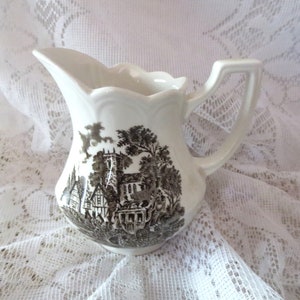 May include: A white ceramic creamer with a brown and black scene of a village with a church and a horse-drawn carriage. The creamer has a scalloped rim and a handle.