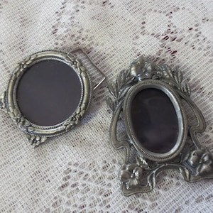 May include: Two antique silver-toned picture frames. One is oval with floral and leaf detailing, the other is round with a decorative border. Both frames have a dark interior and are set against a white lace background.