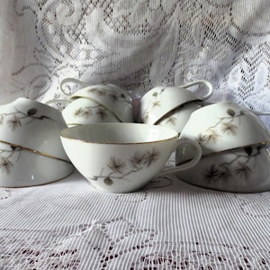 May include: A collection of white porcelain teacups with a delicate gold trim and a painted pine branch design. The teacups are stacked and arranged on a white lace tablecloth, showcasing a vintage aesthetic.