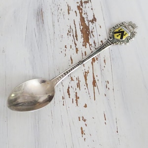 May include: A silver souvenir spoon with a decorative handle and a crest featuring a black and yellow emblem. The spoon's bowl is oval-shaped, and the handle has intricate detailing. The word "OSTENDE" is visible on the crest.
