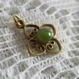 May include: A gold-toned pendant with a four-petal floral design. The center features a round, green gemstone. The pendant has a small loop at the top for a chain. The background is a white, textured fabric.