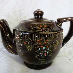 May include: A brown ceramic teapot with a floral design in gold, orange, white, and blue. The teapot has a lid and a handle.