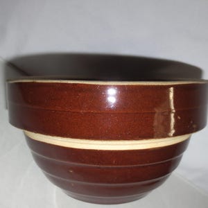 May include: A brown glazed ceramic mixing bowl with a white rim. The bowl has a rounded shape and is made of stoneware.