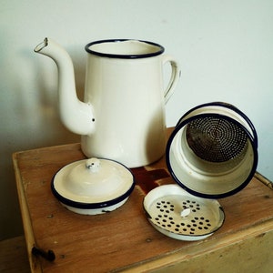 Könnte beinhalten: Weiße Emaille-Kaffeekanne mit blauem Rand und Sieb. Die Kanne steht auf einer Holzkiste mit einem roten Kreuz.