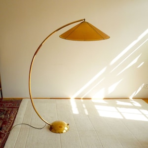 May include: A gold floor lamp with a curved arm and a large, round, tan lampshade. The lamp is on a white rug in a room with hardwood floors.