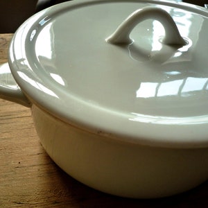 Op de afbeelding: Een witte keramische pot met een deksel. De pot heeft een afgeronde vorm met een handvat aan elke kant en een lusvormig handvat op het deksel. De pot staat op een houten oppervlak.