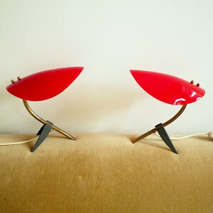 May include: A pair of red table lamps with a curved, gold-tone arm and a black, tripod base. The lamps are on a beige surface.