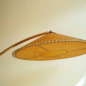 May include: A gold-toned floor lamp with a curved arm and a large, round, tan-colored lampshade with a stitched edge.