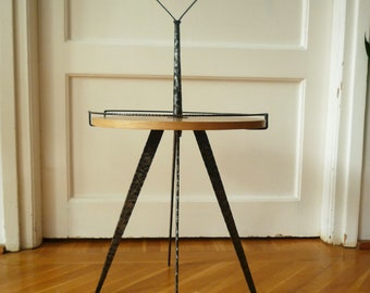 May include: A small, round wooden table with a black metal frame and three legs. The table has a handle on top.