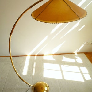 May include: A gold floor lamp with a curved arm and a large, round, tan lampshade. The lampshade has a stitched edge.