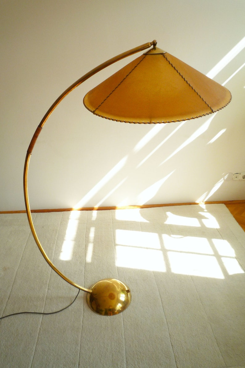May include: A gold floor lamp with a curved arm and a large, round, tan lampshade. The lampshade has a stitched edge.