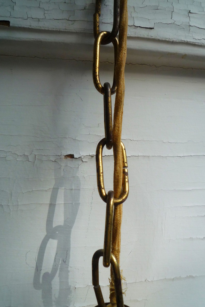 Puede incluir: Una cadena dorada con un cord&oacute;n beige grueso que la atraviesa. La cadena cuelga de un techo blanco con pintura descascarada.