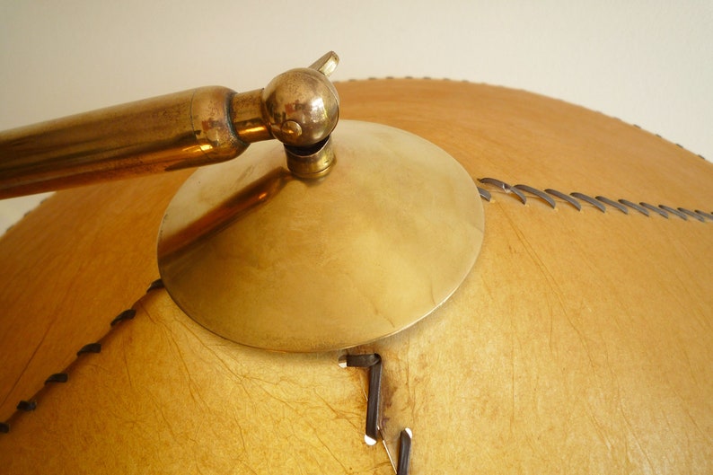 May include: A brass lamp with a large, round, tan leather shade. The shade has a stitched design and a brass trim.