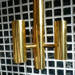 Puede incluir: Una l&aacute;mpara de pared de metal dorado con tres luces. La l&aacute;mpara est&aacute; montada en una pared de azulejos blancos.