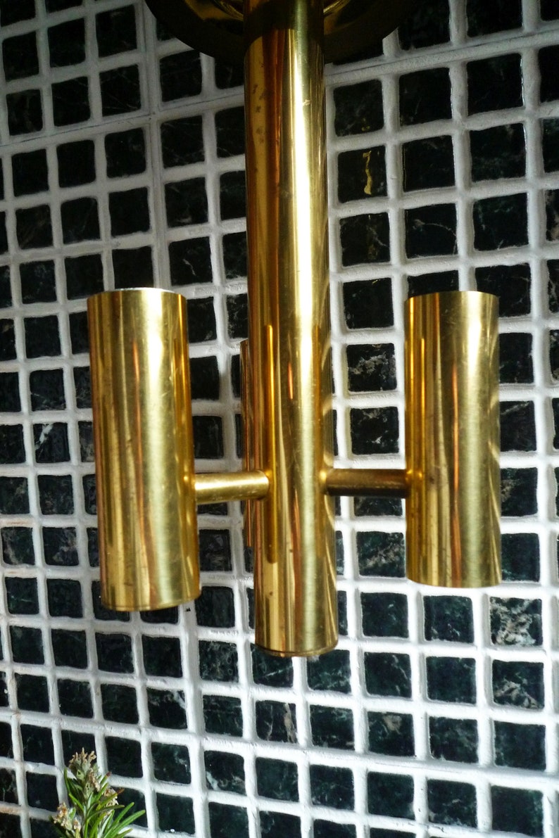 Puede incluir: Una l&aacute;mpara de pared de metal dorado con tres luces. La l&aacute;mpara est&aacute; montada en una pared de azulejos blancos.