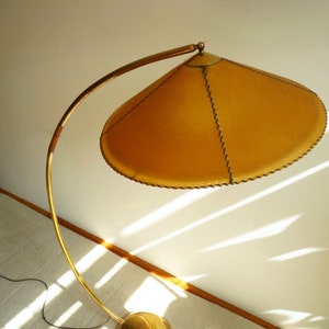 May include: A gold floor lamp with a curved arm and a round, gold base. The lampshade is made of a light brown material and has a stitched edge.