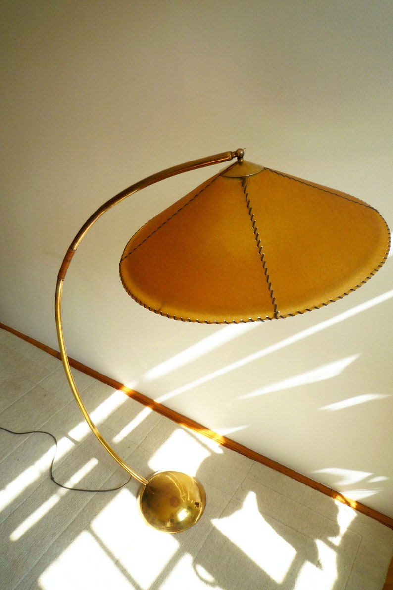 May include: A gold floor lamp with a curved arm and a round, gold base. The lampshade is made of a light brown material and has a stitched edge.