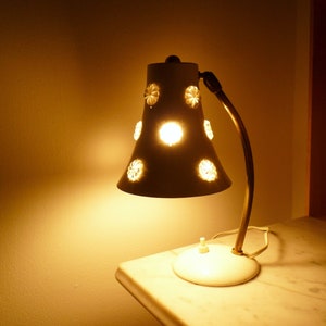 Op de afbeelding: Een witte tafellamp met een witte kap met een bloemenmotief. De lamp heeft een messing arm en voet en wordt verlicht met een warm wit licht.