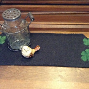 Puede incluir: Una bandeja decorativa de fieltro negro con hojas de roble y bellotas de fieltro verde en cada extremo. Un tarro de cristal transparente con tapa y asa de metal se encuentra en la bandeja, junto con una pequeña figura de pájaro de cerámica.