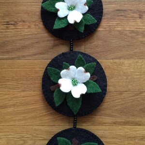 May include: A handcrafted felt decoration featuring three black circular elements, each adorned with a white flower with green and brown accents. The flowers have green centers and are surrounded by green leaves. A black ribbon is attached for hanging.