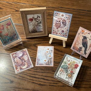 May include: A collection of six framed prints featuring nature-inspired designs. The prints include images of owls, flowers, and a crow. The prints are displayed on a wooden table.