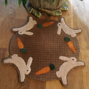 Könnte beinhalten: Ein brauner Filz-Tischset mit gewelltem Rand, mit weißen Hasen-Silhouetten und orangefarbenen Karotten-Applikationen mit grünen Blättern. Das Design ist in einem kreisförmigen Muster angeordnet, ideal für Ostern.